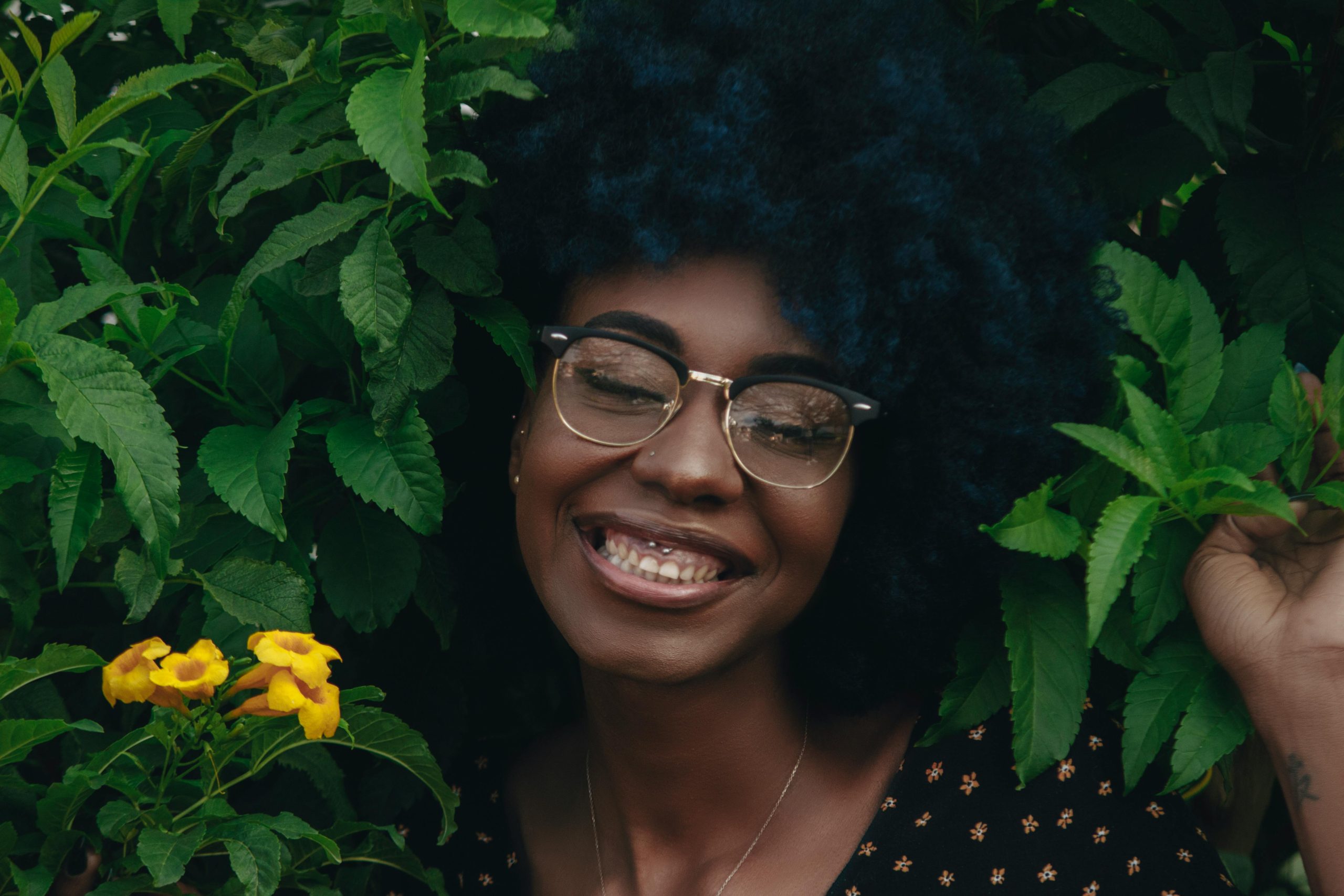 Home A cheerful woman with afro hair grins amidst lush green leaves and yellow flowers outdoors.